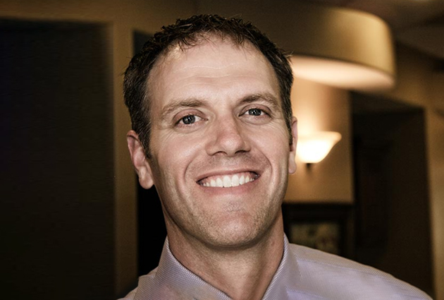 Smiling male professional in light purple shirt, indoor office setting with warm lighting.