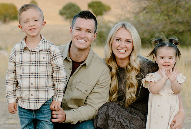 Family of four smiling together outdoors in casual clothing against neutral background.